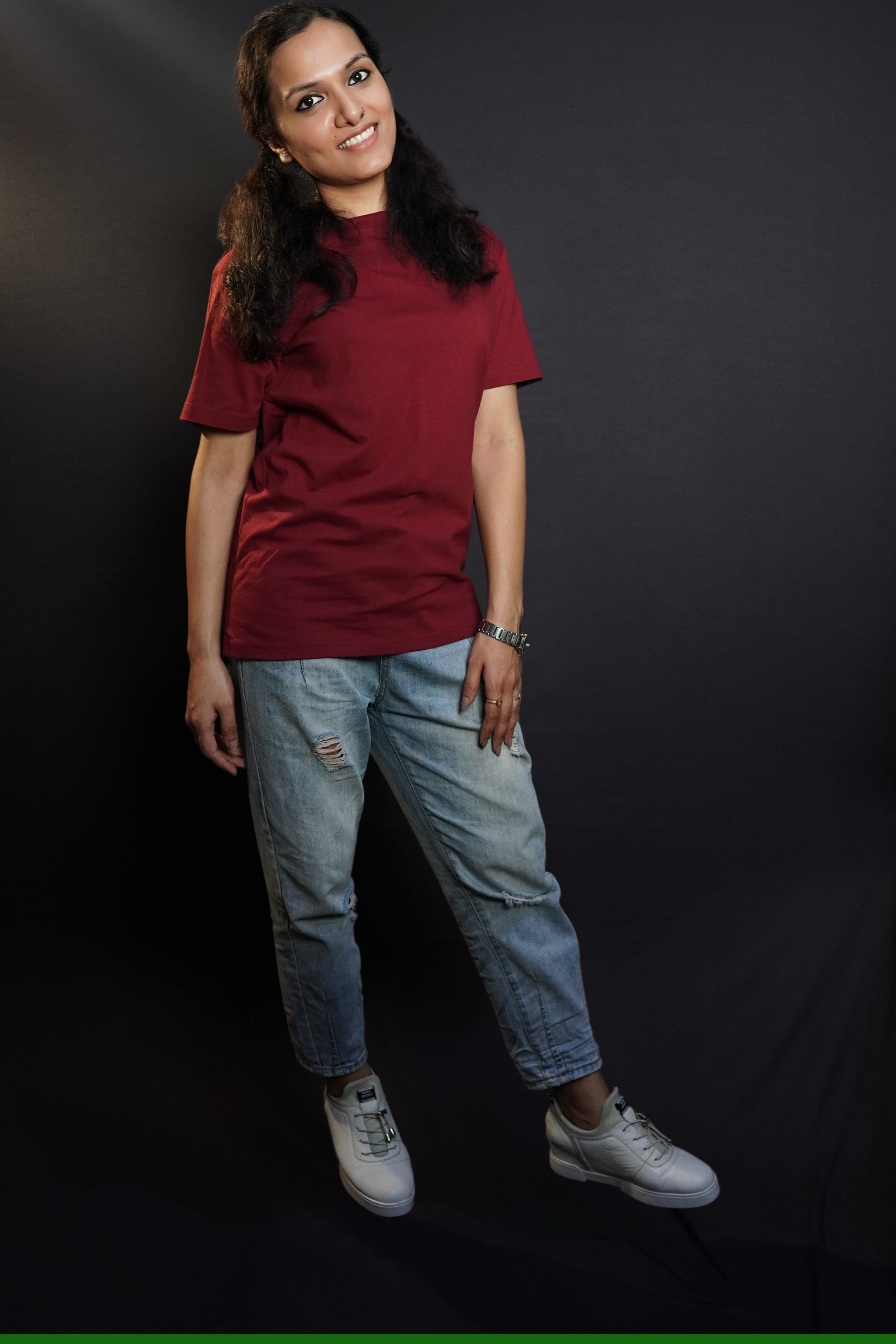 A woman wearing a long length summer cotton rich maroon t-shirt, posing with her hand lying gently on her thigh as she stands against a dark background, showcasing a modish easygoing fit-Muselot