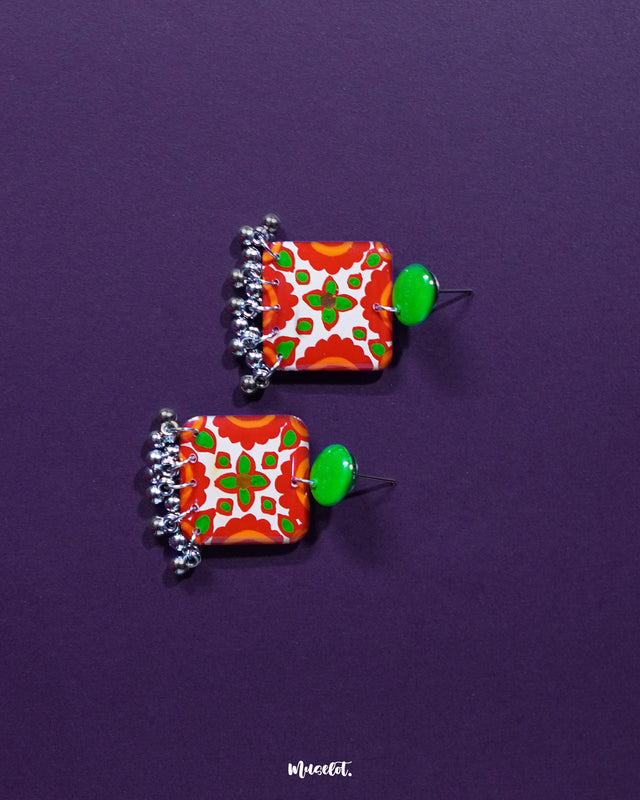 the Zarnigar stud earrings reveals intricate hand-painted floral details in red, green, and ivory set against a metallic base. Dangling silver ghungroos add charm and a traditional rhythm to the design, making them a perfect blend of grace and culture — Muselot.