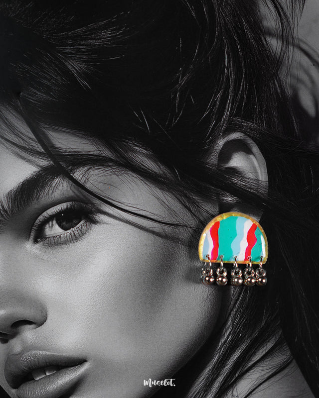 A close-up of a woman wearing the Aabshaar handmade stud earrings, showcasing their vivid wave-patterned design in red, teal, and white, framed with a golden rim and adorned with jingly oxidized silver ghungroos that add movement and charm — Muselot.