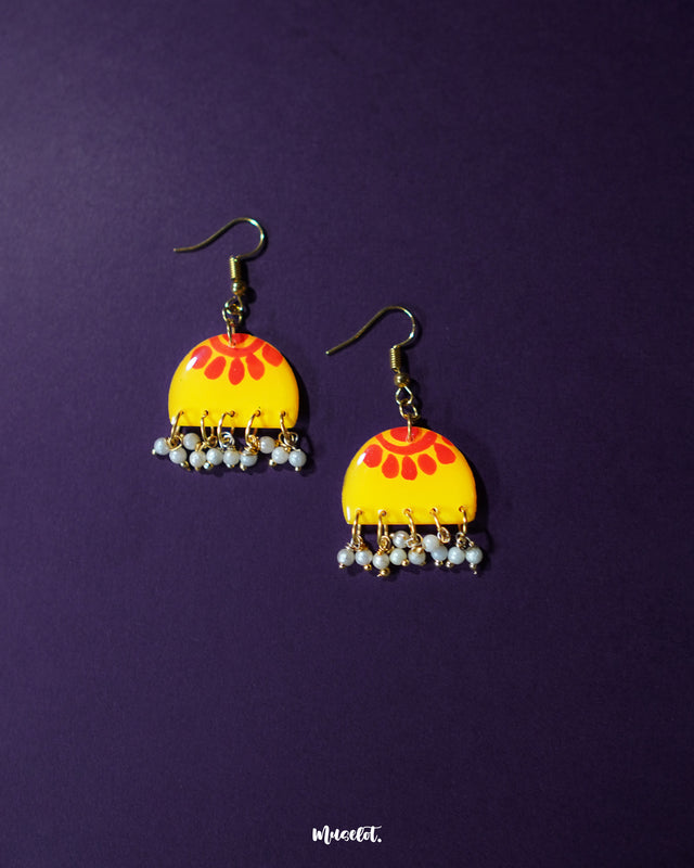 Basantika yellow floral hook earrings displayed on a deep purple background, highlighting their vivid half-moon shape with red floral motifs and delicate blue beaded danglers for a festive touch — Muselot.