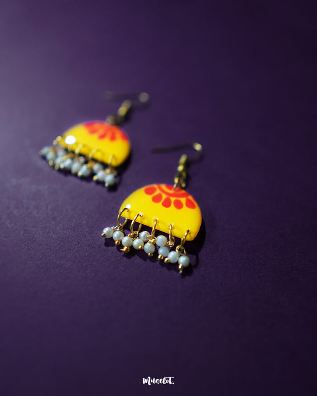 A pair of Basantika yellow floral hook earrings photographed on a dark purple background, showcasing their bold red petal design and dainty blue hanging beads, radiating a bright, festive charm — Muselot.
