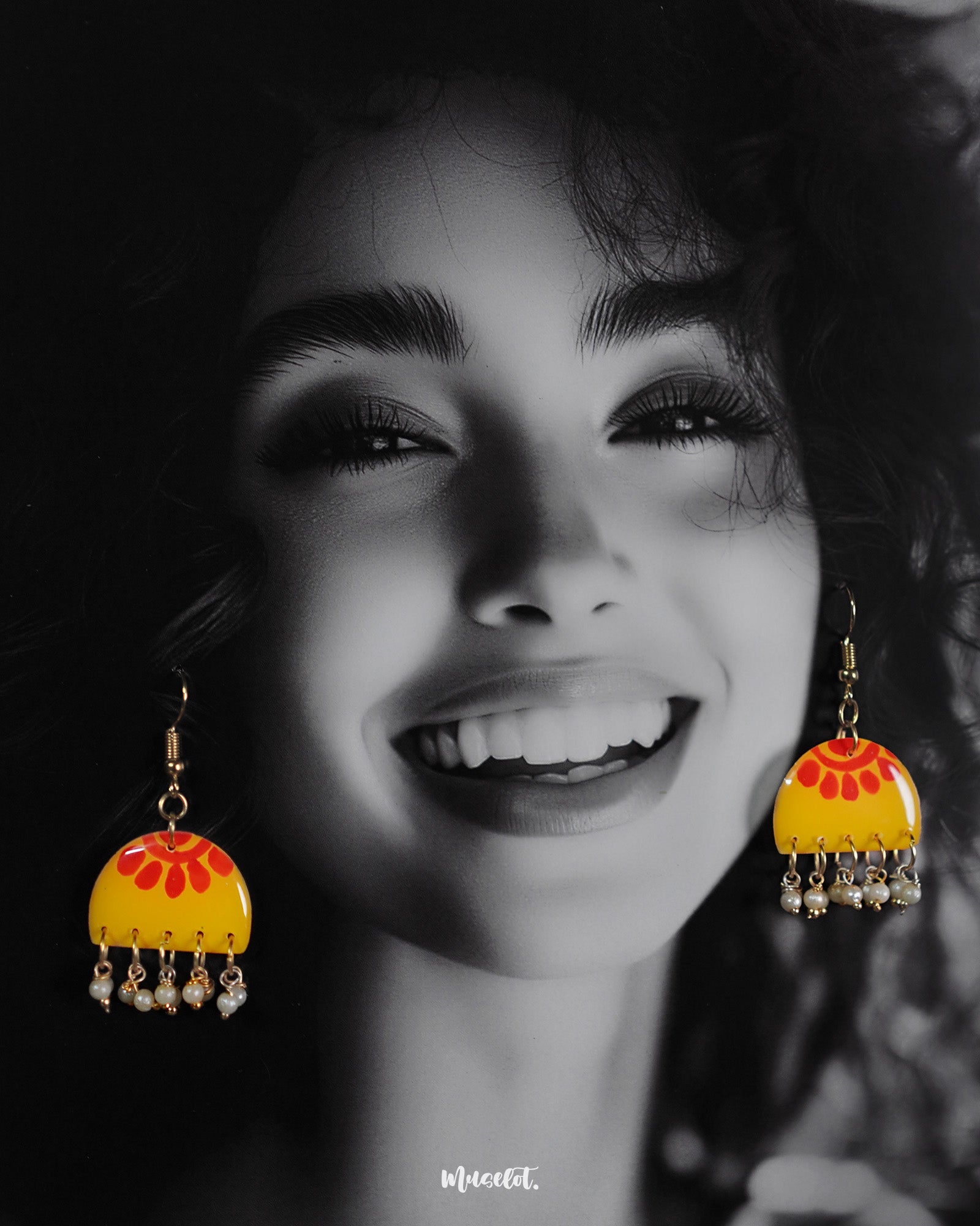 A woman wears the Basantika yellow and red hook earrings, highlighting the hand-painted semicircle motif in warm hues and delicate blue bead danglers that add a touch of artisanal flair — Muselot.