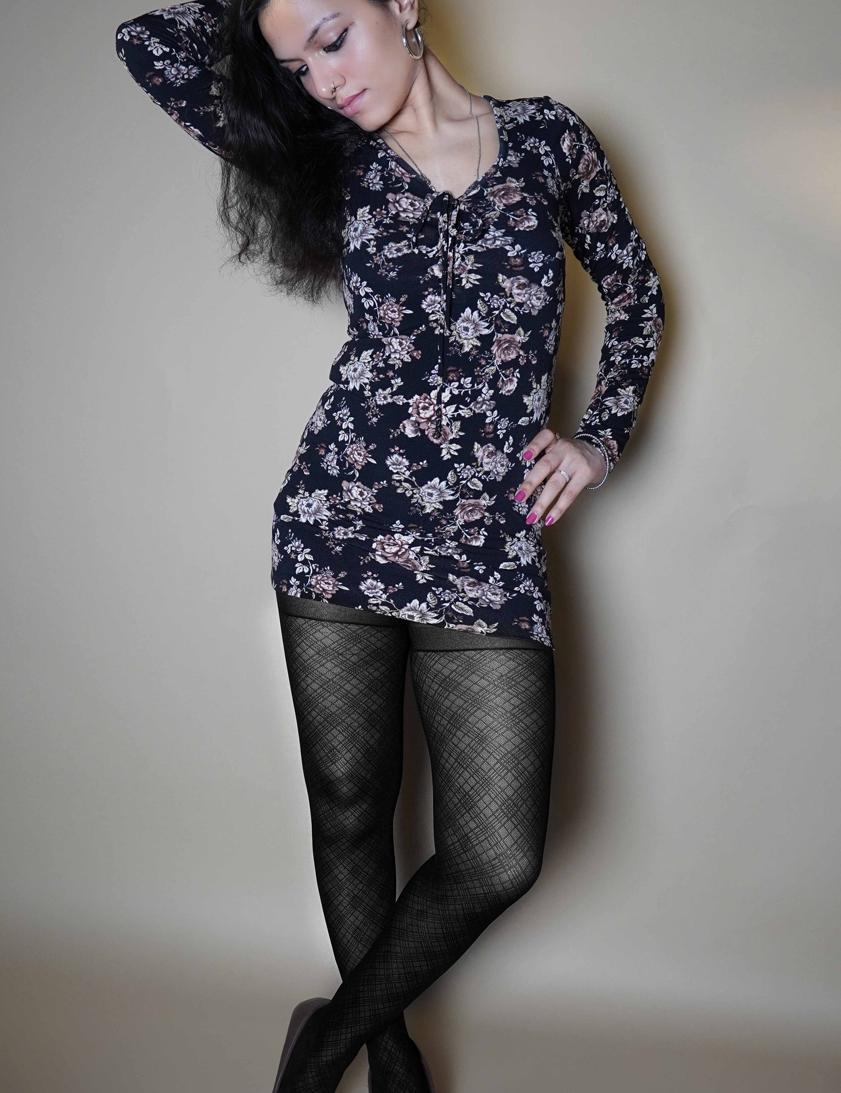 A woman wearing black transparent sheer patterned stockings, posing with her hand on her waist and other behind her head as it is bent sideways, standing cross legged, highlighting the relaxed modern look-Muselot