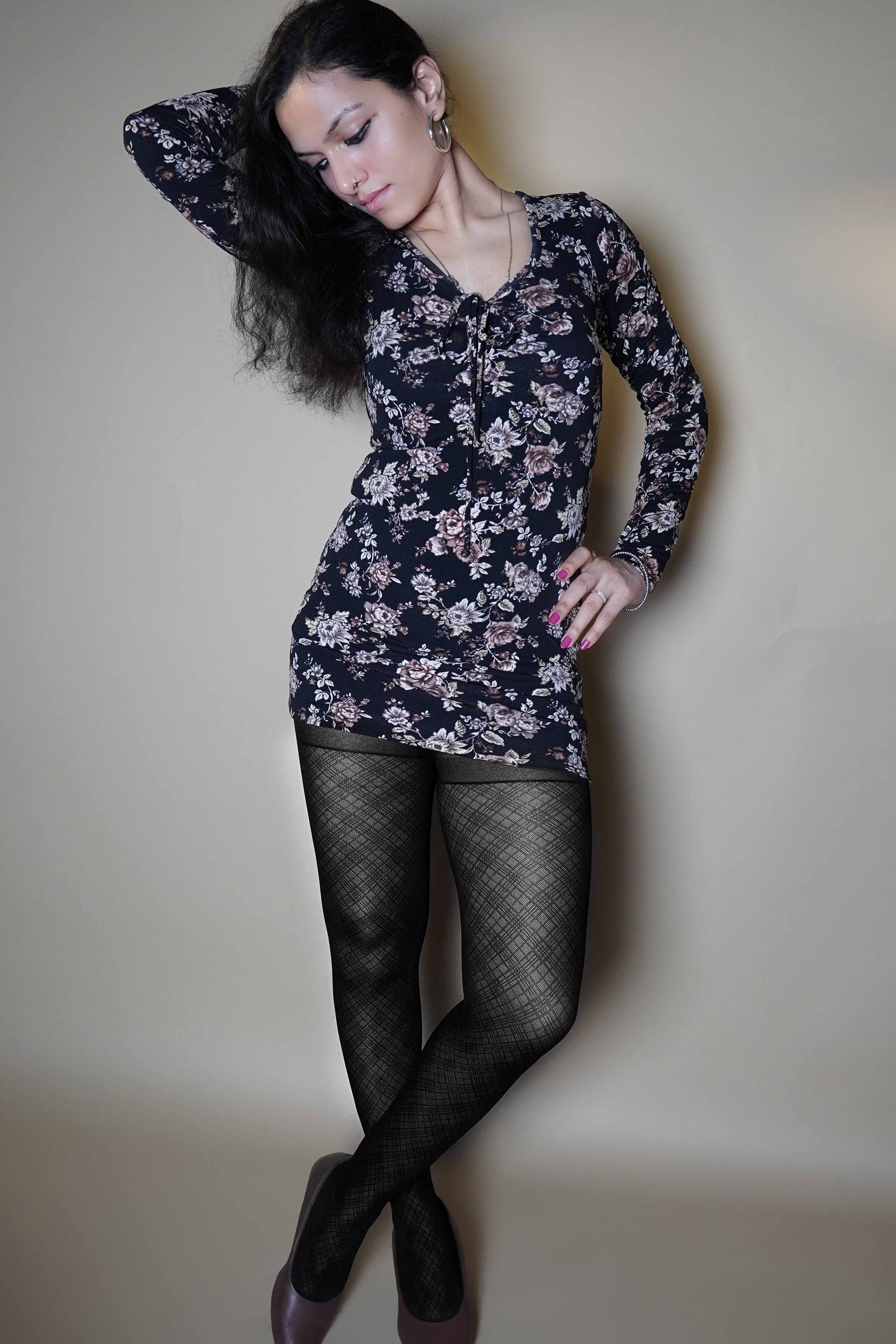 A woman wearing black transparent sheer patterned stockings, posing with her hand on her waist and other behind her head as it is bent sideways, standing cross legged, highlighting the relaxed modern look-Muselot