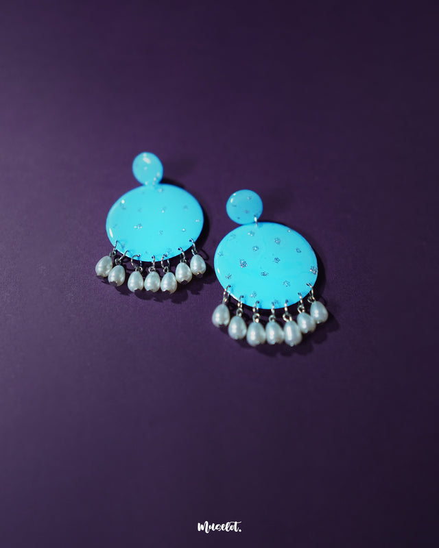 Blue pearl earrings featuring small and large round turquoise blue discs decorated with delicate silver glitter accents, complemented by dangling white pearl-like beads, elegantly showcased to highlight their vibrant and festive design – Muselot.