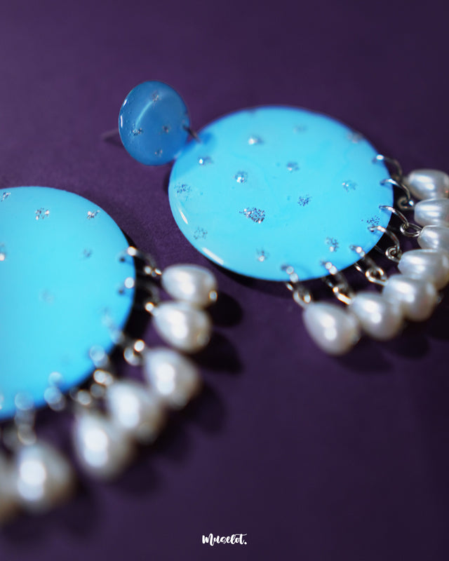 Close-up of the Blue pearl earrings, showcasing small and large round turquoise blue discs with fine silver glitter accents, finished with dangling white pearl-like beads, highlighting their intricate detailing and elegant charm – Muselot.