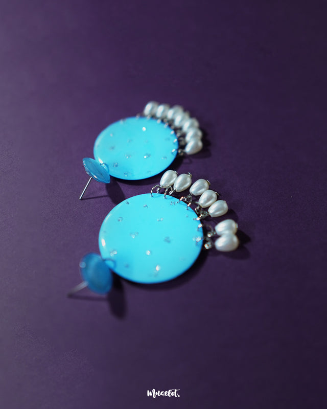 Blue pearl earrings featuring layered small and large turquoise blue discs with subtle silver glitter accents, adorned with dangling white pearl-like beads, beautifully displayed to showcase their vibrant color and delicate craftsmanship – Muselot.