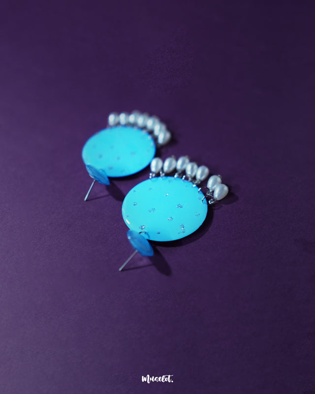 Blue pearl earrings featuring small and large turquoise blue discs with subtle silver glitter accents, complemented by dangling white pearl-like beads, artistically showcased to emphasize their vibrant color and elegant detailing – Muselot.