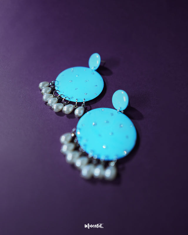 Blue pearl earrings featuring small and large round turquoise blue discs with subtle silver glitter accents, paired with dangling white pearl-like beads, elegantly displayed to highlight their vibrant and festive charm – Muselot.