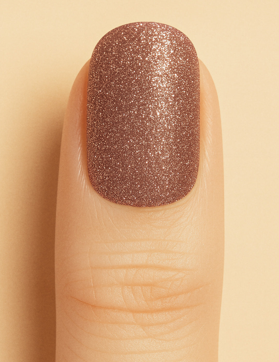 A close-up of a model’s finger wearing Caramel Glow Glitter nail stickies against a beige background, showcasing the warm caramel-brown shade with shimmering glitter, highlighting its radiant glossy finish – Muselot.