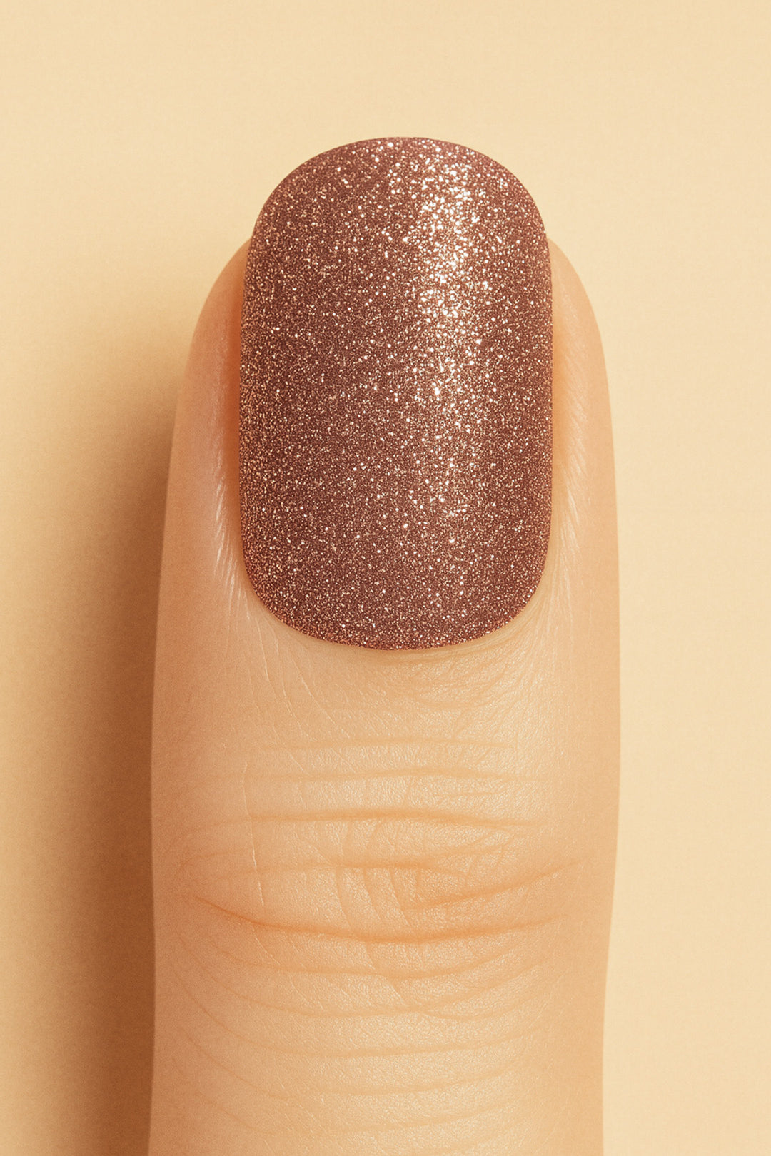 A close-up of a model’s finger wearing Caramel Glow Glitter nail stickies against a beige background, showcasing the warm caramel-brown shade with shimmering glitter, highlighting its radiant glossy finish – Muselot.
