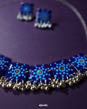 Falakzaar blue necklace and earring set displayed against a purple background, showcasing the hand-painted detailing and highlighting the elegant craftsmanship and vibrant charm of this statement set — Muselot.