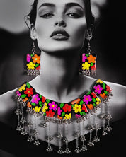A model wearing the floral jewelry set featuring a bold necklace and matching earrings. The black base is beautifully hand-painted with bright, multicolored flowers, and finished with silver gungroo chains that dangle playfully. The vibrant design stands out against the outfit, adding a joyful, boho charm to the overall look — Muselot.