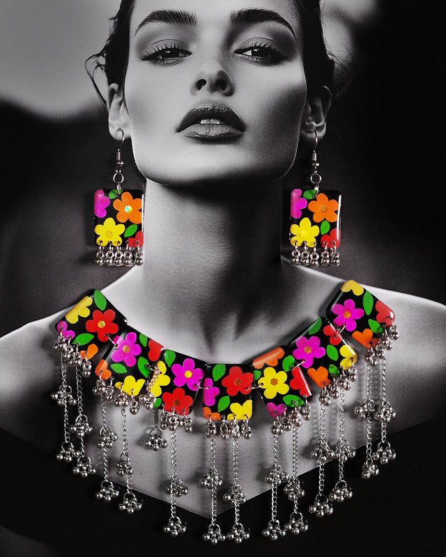 A model wearing the floral jewelry set featuring a bold necklace and matching earrings. The black base is beautifully hand-painted with bright, multicolored flowers, and finished with silver gungroo chains that dangle playfully. The vibrant design stands out against the outfit, adding a joyful, boho charm to the overall look — Muselot.