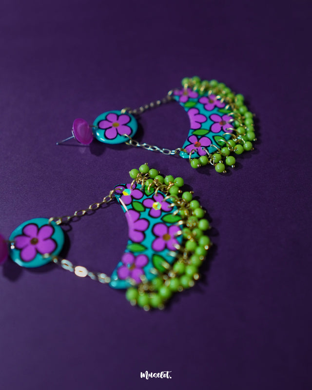 A pair of Gulbahaar chandbali earrings laid flat, showcasing the crescent shape adorned with purple and green floral artwork and dangling yellow beads, highlighting their artisanal charm — Muselot.