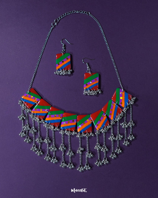 Gulrang multicolor resin jewelry set featuring a silver chain necklace with rectangular charms in bold red, green, blue, and purple diagonal stripes accented with dots, paired with matching earrings and intricate silver bead tassels, beautifully displayed on a deep purple background – Muselot.