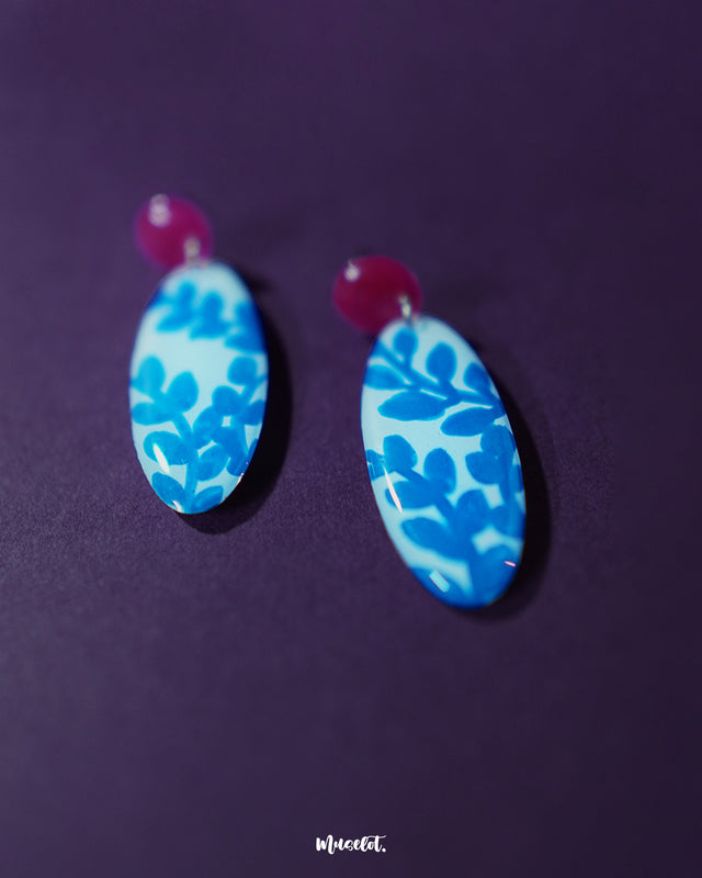 Close-up of the Gulsaba blue handmade drop earrings, showcasing glossy red circular studs paired with elongated white oval drops detailed with bold hand-painted blue leafy patterns, highlighting the intricate brushwork and vibrant contrast – Muselot.