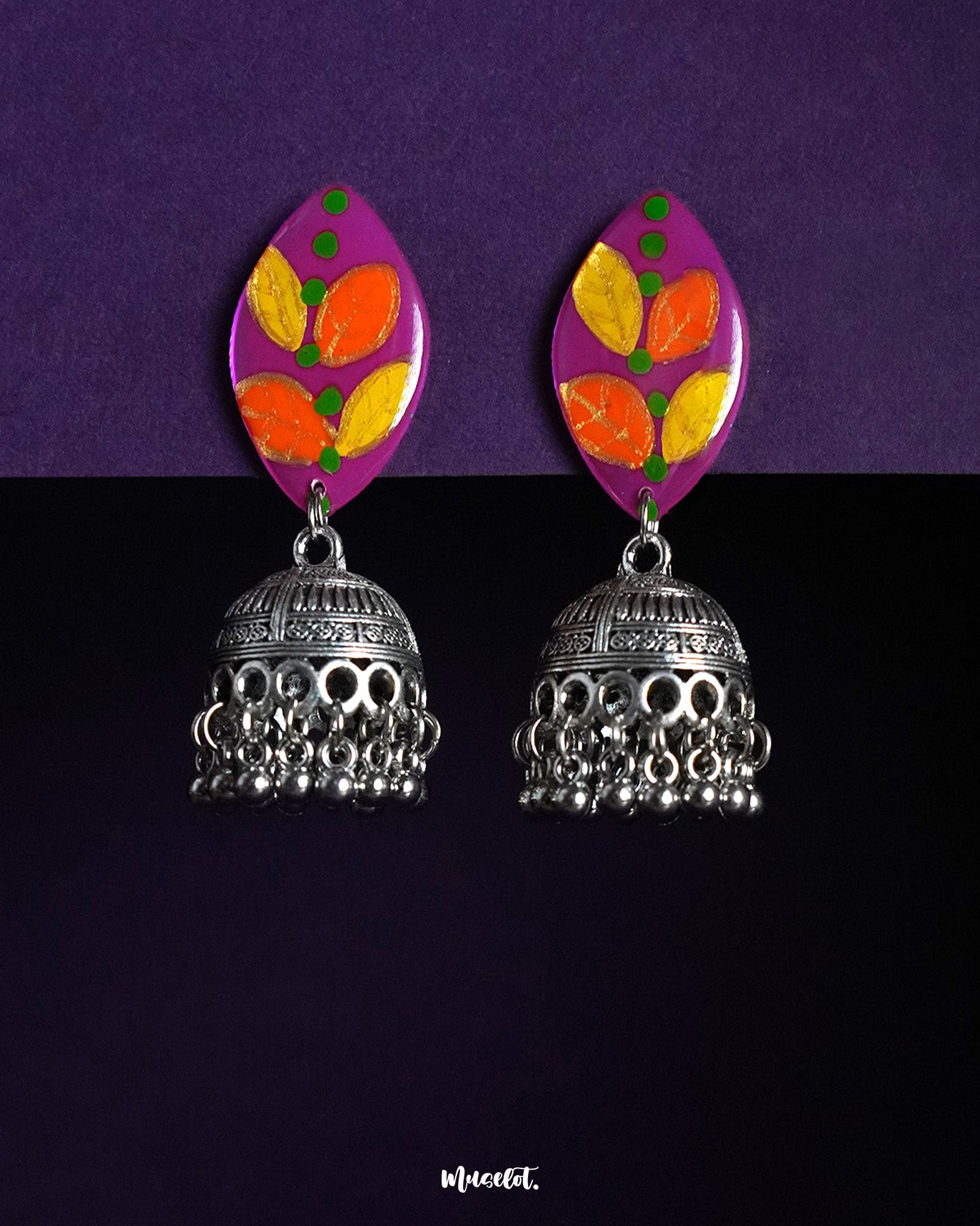 Gulzarish jhumkas featuring marquise-shaped tops painted in vibrant purple with orange and yellow leaf motifs accented by green dots, paired with intricately carved oxidized silver dome-shaped jhumka drops adorned with delicate hanging beads, beautifully displayed on a deep purple background – Muselot.