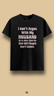 Black graphic t-shirt showcasing the statement “I Can’t Argue With My Husband He Is Older Than Me And Old People Don’t Listen.” in soft high-contrast lettering, highlighting witty relationship humor in a clean beige studio presentation – Muselot
