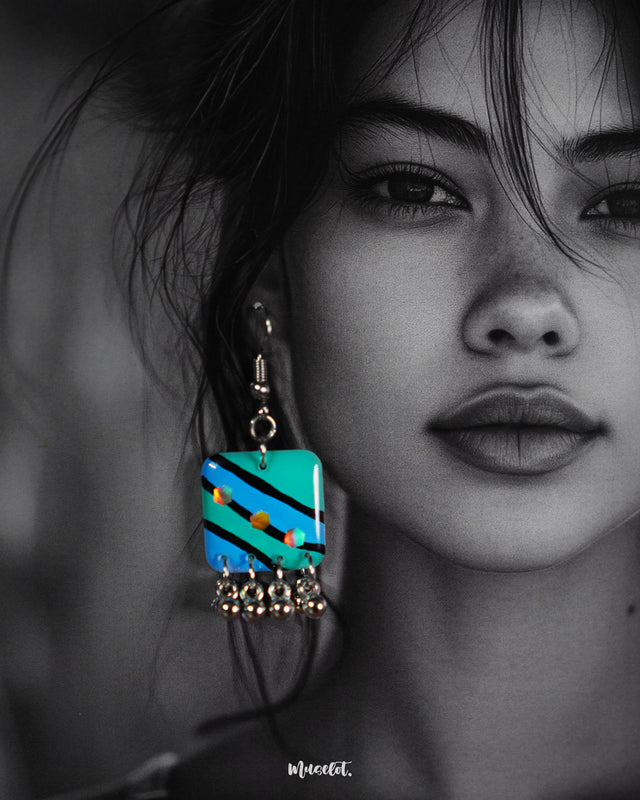 A woman wears the Jaltarang strip drop earrings, their hand-painted green and blue stripes standing out vividly against her skin, while the silver ghungroos at the bottom add a rhythmic charm to her look—Muselot.