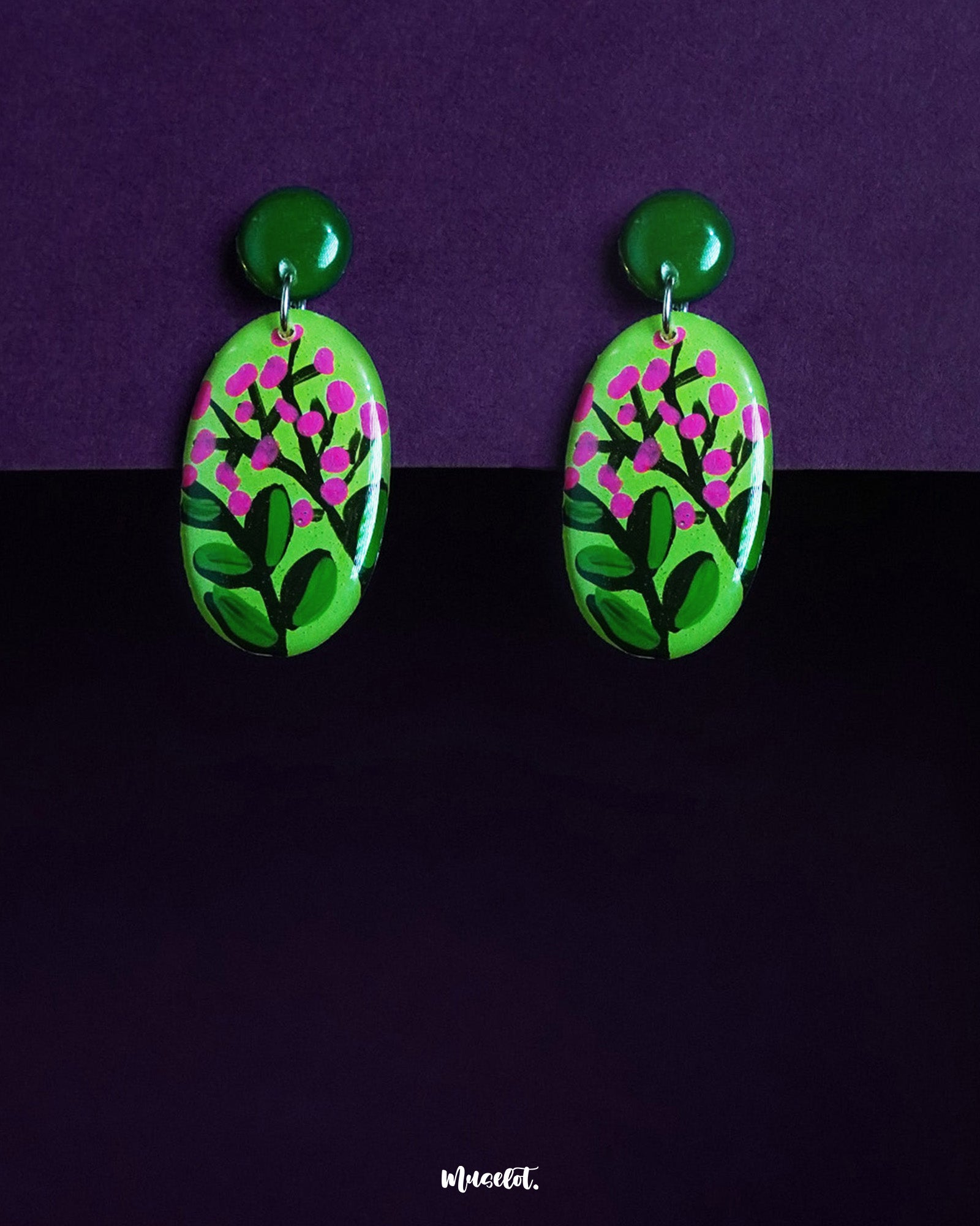 The Kalibaan drop earrings are showcased against a deep purple background, featuring a vivid green oval base hand-painted with delicate pink floral motifs,highlighting the bold charm of nature-inspired design, the earrings are topped with glossy round studs that add a playful yet grounded touch — Muselot.