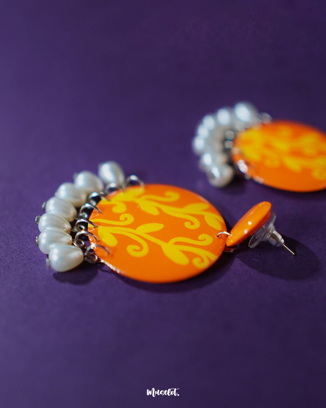 A close-up of the Kesarbaan orange and yellow drop earrings revealing the intricate brushstrokes of the floral design, paired with elegant white pearl drops that add charm to their festive handcrafted appeal — Muselot.