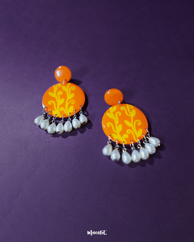 Kesarbaan orange‑and‑yellow drop earrings laid flat against a soft background, highlighting the vivid hand‑painted sunburst motif and showcasing the graceful white pearl drops that add festive elegance — Muselot.