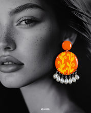 A woman wearing the Kesarbaan pearl drop earrings, where the vibrant orange discs with yellow folk motifs draw the eye, while the dangling white pearls softly frame her jawline, adding a graceful charm to her look – Muselot.