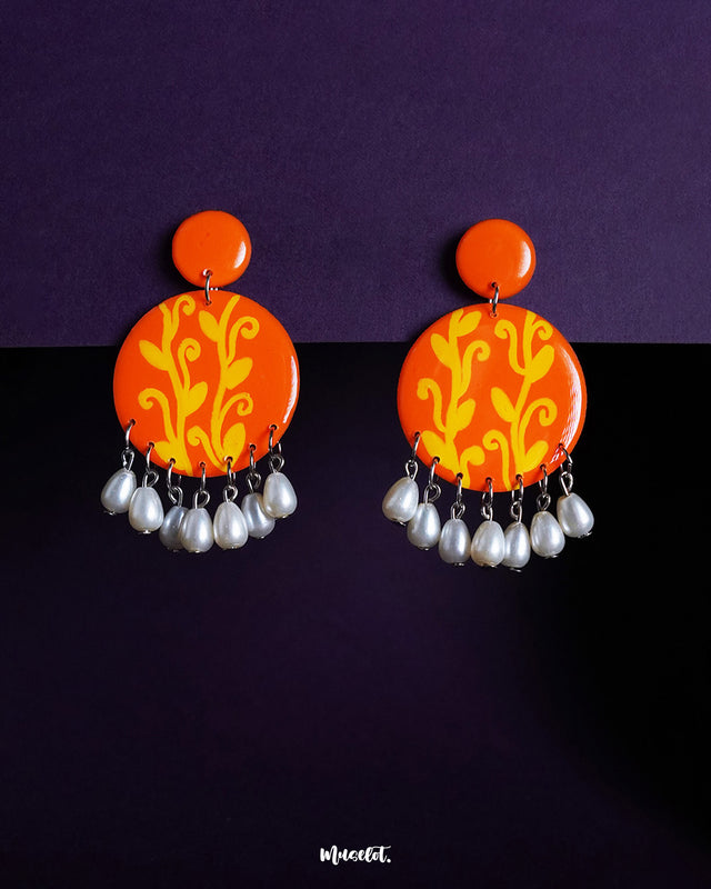 Kesarbaan pearl drop earrings set against a deep purple backdrop, featuring vibrant orange discs adorned with yellow folk motifs and finished with delicate white pearl drops that add a touch of festive elegance – Muselot.