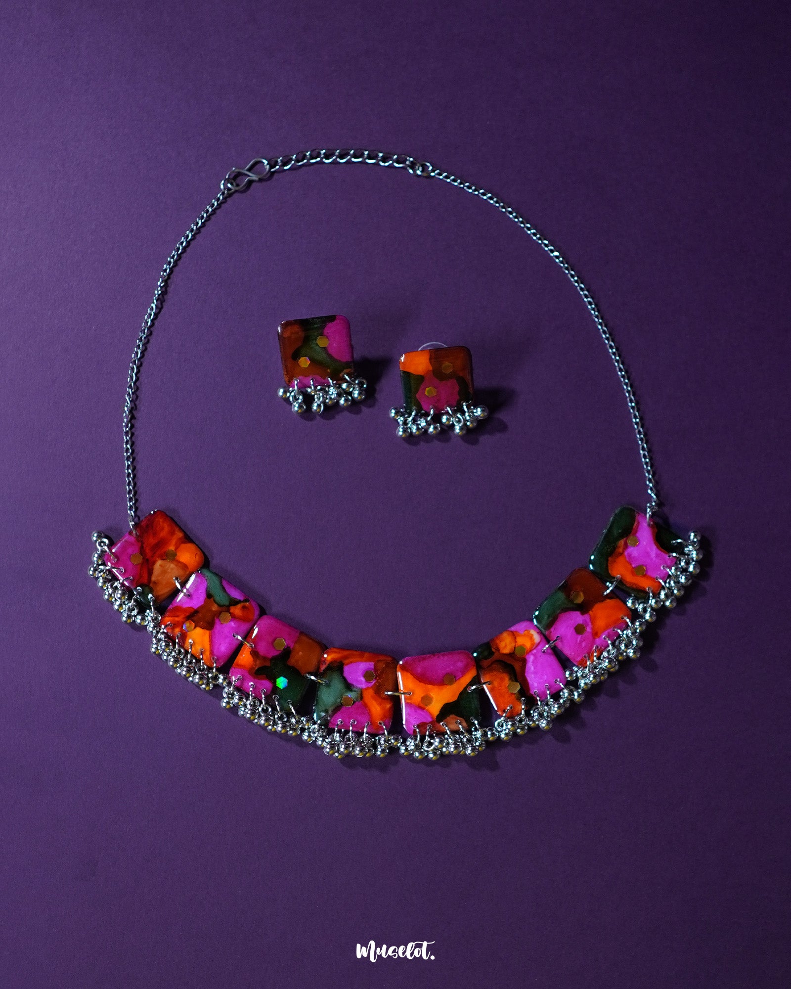 Khushk Gul handmade floral necklace set featuring a silver chain necklace and matching square stud earrings, adorned with vibrant pink, orange, and green abstract floral patterns and delicate silver bead accents, beautifully showcased on a deep purple background – Muselot