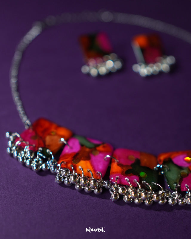 Zoomed-in shot of the Khushk Gul handmade floral necklace set on a purple background, featuring vivid pink, orange, and green abstract floral-patterned square charms with silver bead accents, capturing the fine details and rich colors – Muselot.