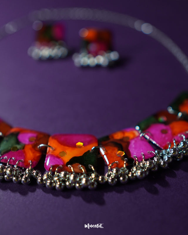 Khushk Gul handmade floral necklace set with a silver chain and matching square stud earrings, adorned with vibrant pink, orange, and green abstract floral patterns and silver bead accents, elegantly displayed on a deep purple background – Muselot.