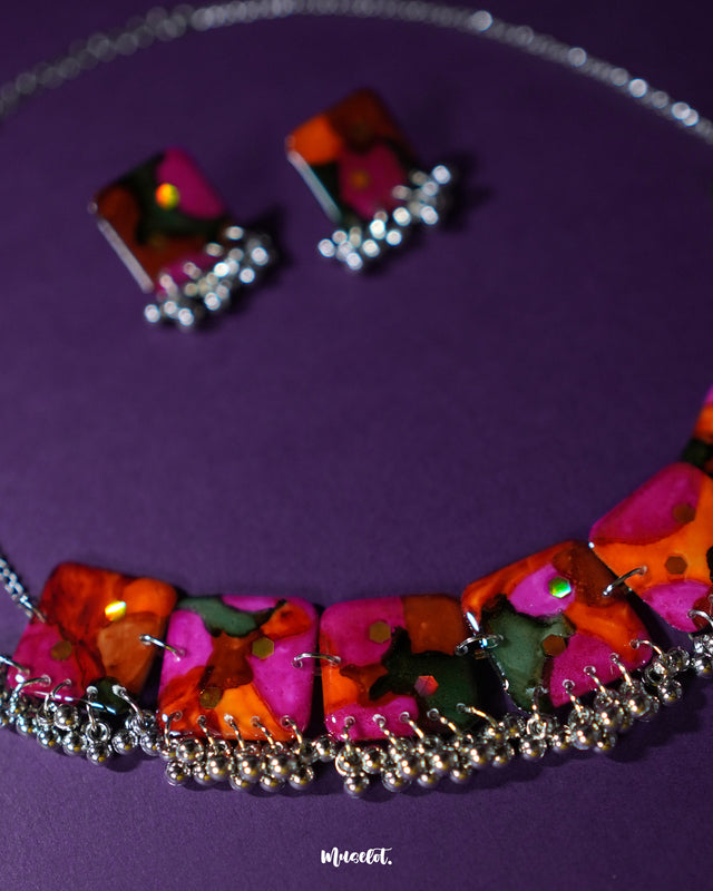 Khushk Gul handmade floral necklace set featuring a silver chain and matching square stud earrings, decorated with vivid pink, orange, and green abstract floral patterns and delicate silver bead accents, artistically showcased on a textured deep purple background – Muselot.
