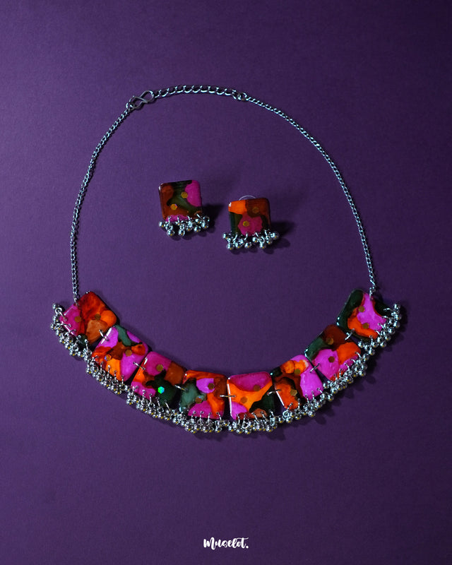 Khushk Gul handmade floral necklace set featuring a silver chain necklace and matching square stud earrings, adorned with vibrant pink, orange, and green abstract floral patterns and delicate silver bead accents, beautifully showcased on a deep purple background – Muselot