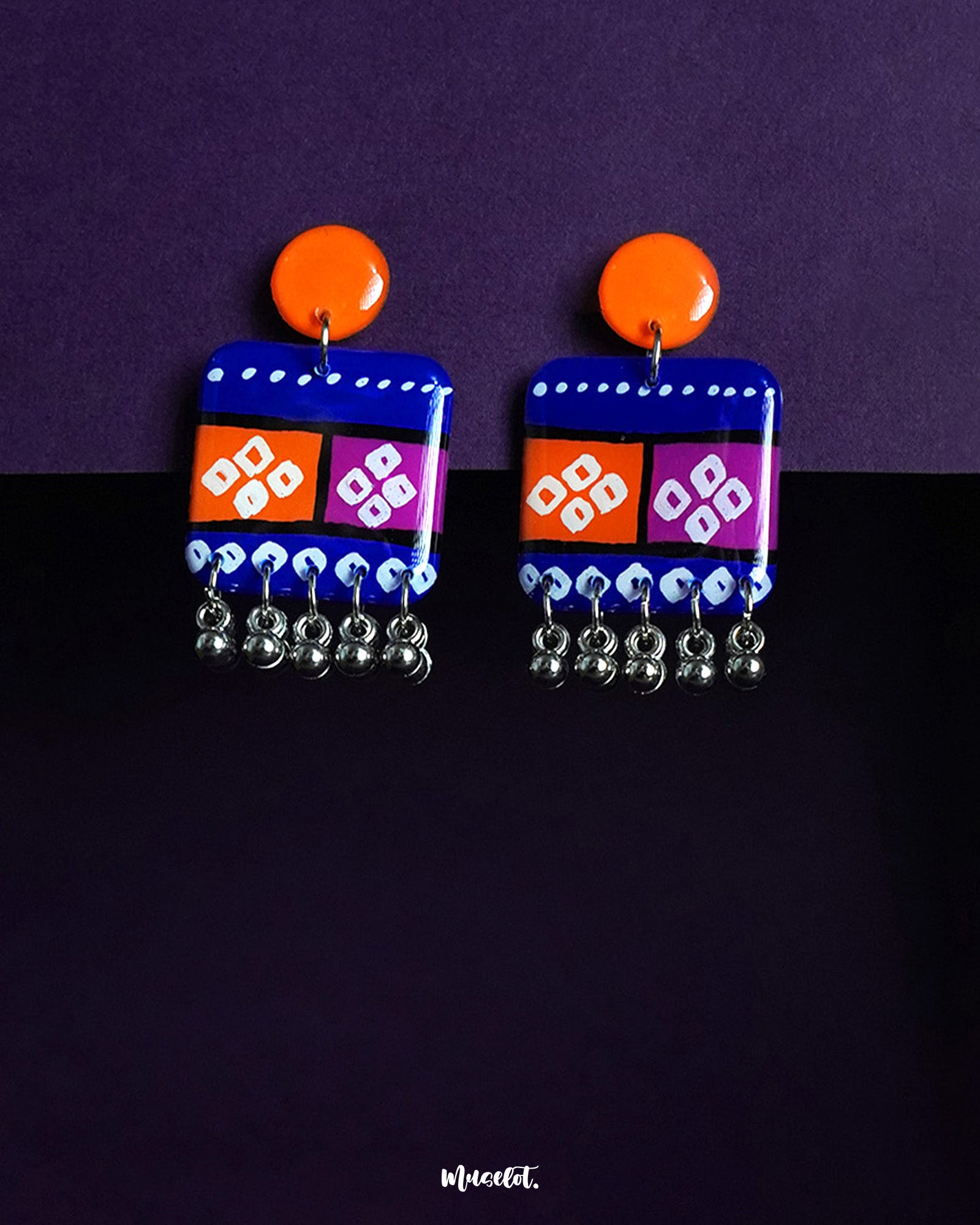 Leheriya boho earrings featuring bright orange round studs connected to rectangular drops painted in vibrant blue with orange and magenta blocks, adorned with white geometric patterns and dotted detailing, finished with dangling silver beads, beautifully displayed against a deep purple background – Muselot.