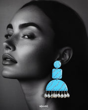 A model wearing the Lehrbaan drop earrings showcases their vibrant sky blue color and unique geometric design. The dangling white pearls add graceful movement, while the glittery silver waves catch the light, making them a standout accessory for festive or artistic looks — Muselot.