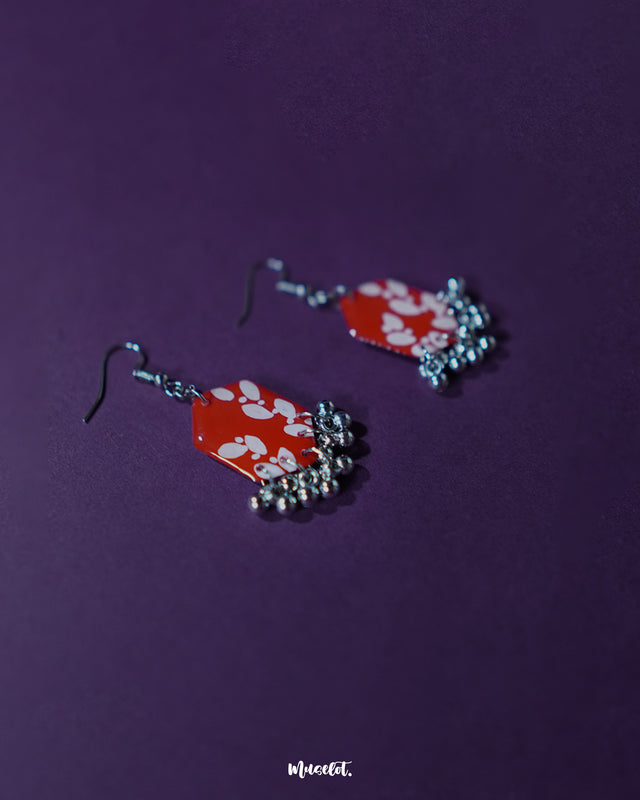 A Mehar red floral hook earrings from Muselot placed against a purple background — showcasing vivid red hexagonal drops with delicate white floral motifs and metallic ghungroo accents, highlighting the intricate handcrafted design and vibrant color contrast.
