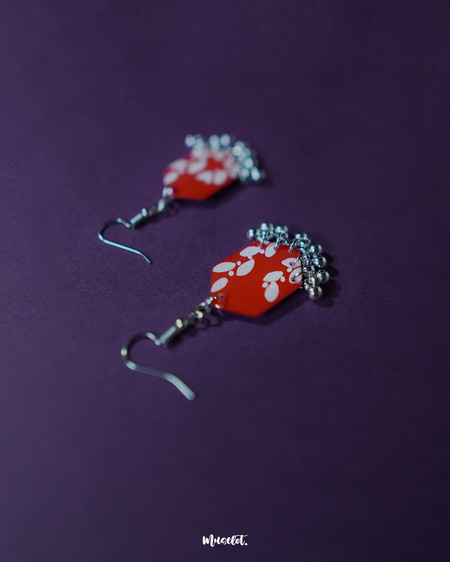 The Mehar red floral hook earrings from Muselot displayed at an angle, showcasing the intricate white floral motifs on bold red hexagonal drops, with metallic ghungroo accents highlighting the detailed craftsmanship and artistic flair.
