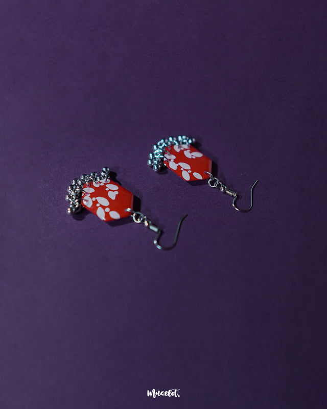 The Mehar red floral hook earrings from Muselot laid flat — highlighting the symmetrical hexagonal shape, vivid red enamel, delicate white floral detailing, and metallic ghungroo accents that showcase the piece’s handcrafted charm and cultural finesse.