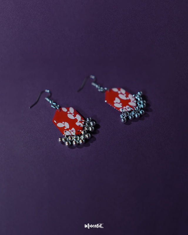 The Mehar red floral hook earrings from Muselot captured from a side angle — showcasing the dimensional depth of the hexagonal drops, the intricate white floral artwork, and the subtle sway of metallic ghungroos that lend movement and grace to the piece.