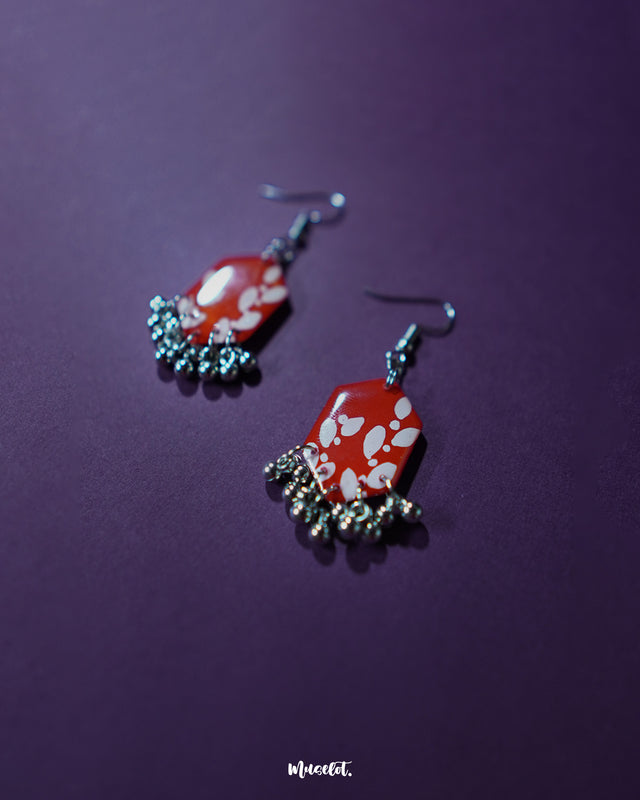 The Mehar red floral hook earrings displayed with a soft shadow play ,showcasing the intricate hand-painted floral motifs on rich red enamel and the gentle shimmer of metallic ghungroos, highlighting the earrings’ artistic depth and elegance.