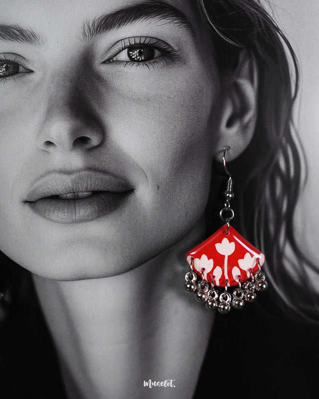 A close-up of a model wearing the Meherbaan red floral earrings, highlighting the glossy red base with hand-painted white tulip designs and jingling silver ghungroo drops that gently sway, adding a graceful festive accent — Muselot.