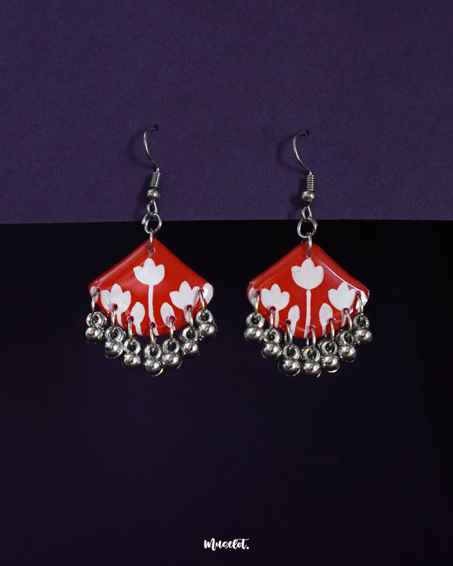 A pair of handcrafted red floral earrings with white tulip motifs on a glossy red base, accented by delicate silver ghungroo-style danglers. The vibrant design and playful jingles give it a festive, artisanal charm — Muselot.