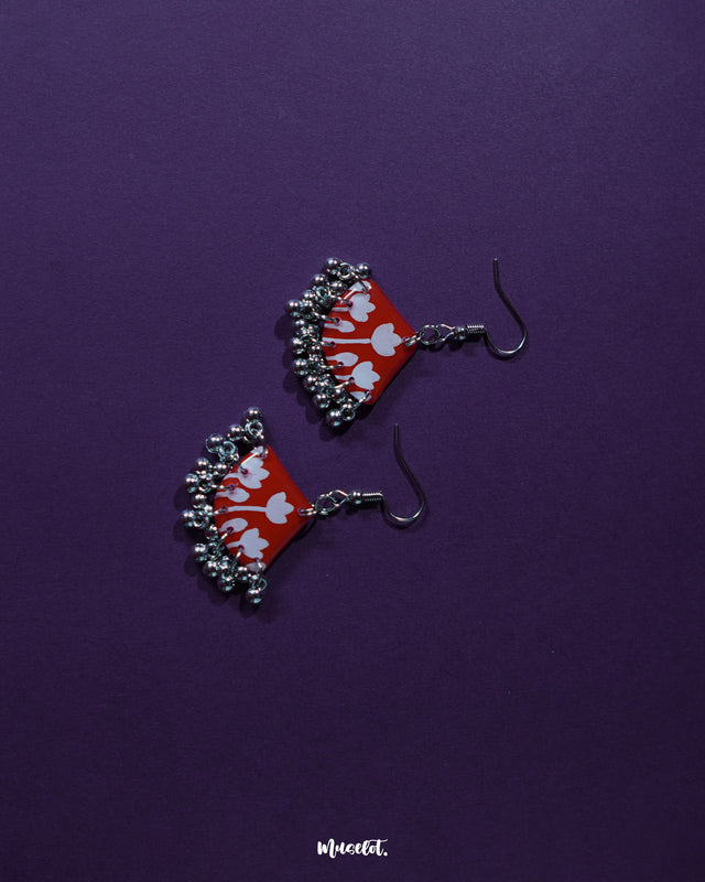 Meherbaan red floral hook earrings featuring a hand-painted tulip motif on a rich red base, adorned with delicate silver ghungroo charms, set against a deep purple backdrop — Muselot.