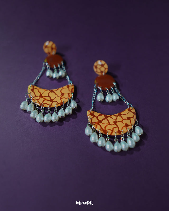 Mittika chandbali earrings laid flat against a textured neutral background, highlighting their crescent shape, floral center motif, and delicate pearl hangings for a graceful ethnic charm — Muselot.