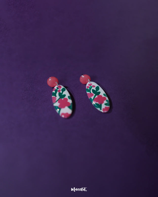 A pair of Nargis pink floral drop earrings resting flat, showcasing the oval-shaped design with vivid pink flowers and green leaves on a white base, highlighting the glossy enamel and handmade details — Muselot.
