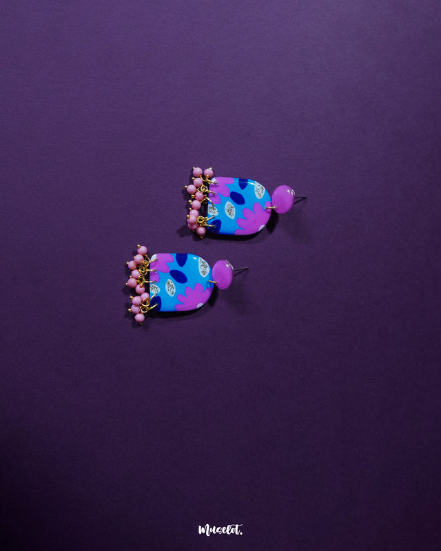 Neelzair purple floral earrings placed against a rich purple background, highlighting the glossy surface, hand-painted floral patterns in pink and blue, and showcasing the dangling pink beads that add a lively touch — Muselot.