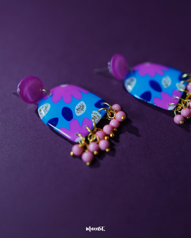 The Neelzair purple floral earrings rest against a purple background, highlighting their vivid color palette and intricate floral design with beaded fringes — Muselot.
