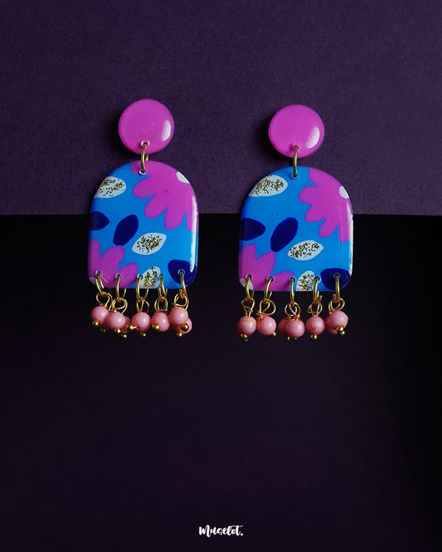Neelzair purple floral earrings with pink stud tops and dangling pink beads, showcasing vibrant hand-painted petals and a rich contrast of blue and fuchsia tones against a deep purple background — Muselot.