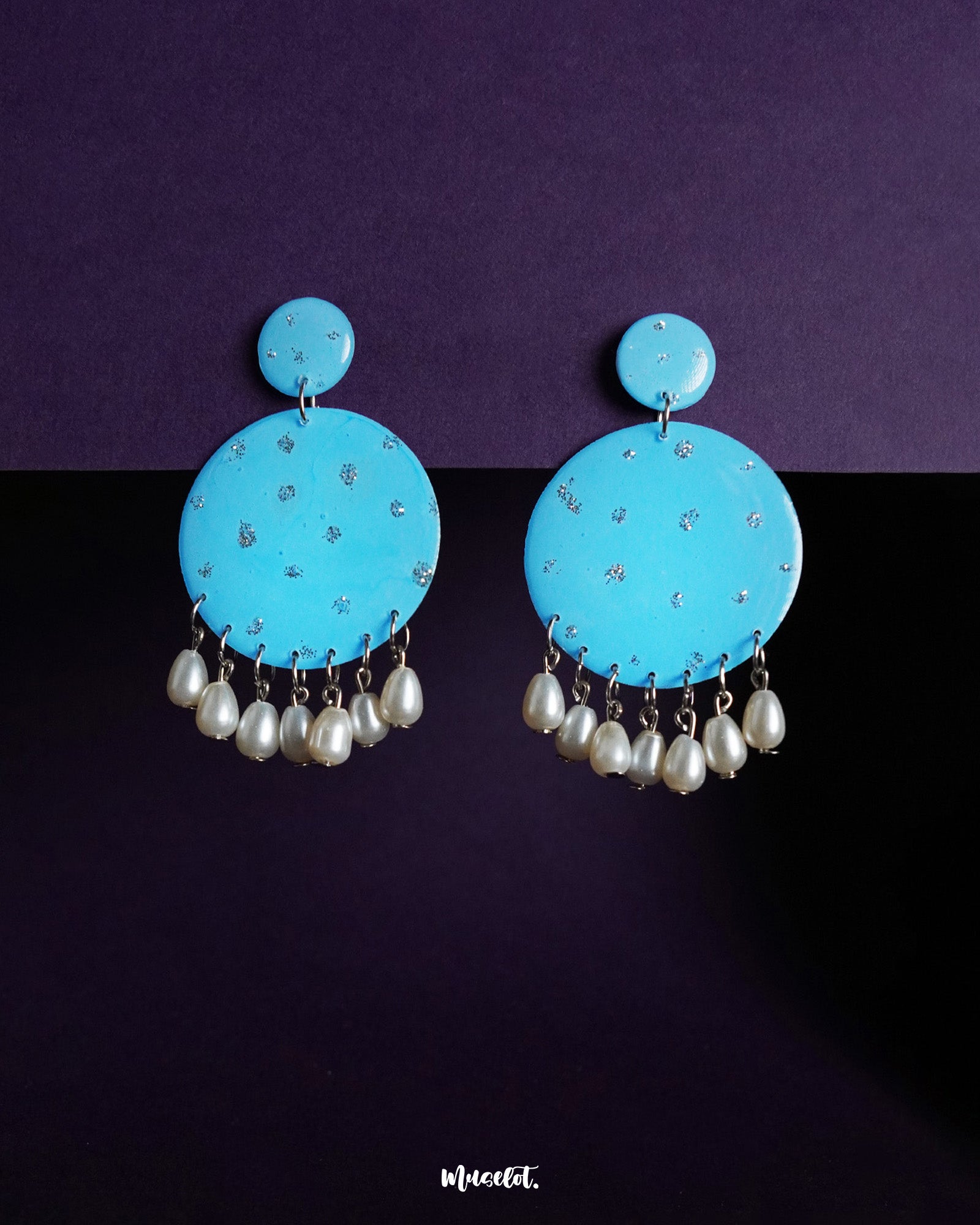 Neerangi pearl earrings featuring small and large round turquoise blue discs embellished with subtle silver glitter accents, finished with dangling white pearl-like beads, beautifully displayed against a deep purple background – Muselot.